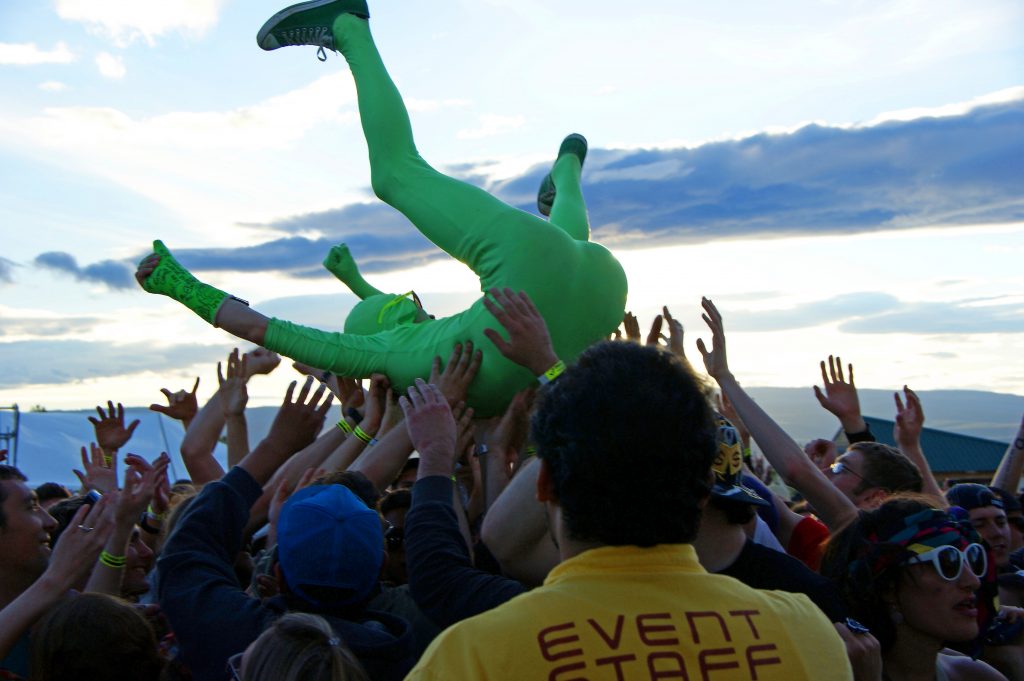 A person in a neon green full body "MorphSuit" crowd surfs above people at an outdoor music event, whilst a member of event staff looks on